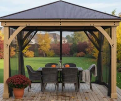 Wooden gazebo with mesh curtains and a metal roof on a deck, featuring a dining table with pumpkins, a vase, and wicker chairs, set in a backyard with autumn foliage and residential houses in the background.