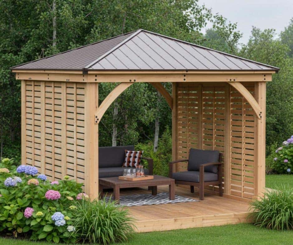 Wooden gazebo with a metal gabled roof and slatted walls on three sides, featuring cushioned armchairs and a coffee table with glasses and a bottle, set on a patterned rug in a landscaped garden with hydrangeas and tall trees.