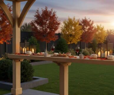 Outdoor autumn dining setup under a wooden pergola, with white pumpkins, lanterns, and fall leaves decorating the table, surrounded by vibrant foliage and a pink-hued sky.