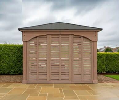 Costco Grand Gazebo 12ft Privacy Wall Costco Accessories Premium Cedar Gazebos with Aluminium Roofs Wooden gazebo with a dark pitched roof and slatted front design, set on a stone-tiled patio with a green hedge and residential houses in the background.
