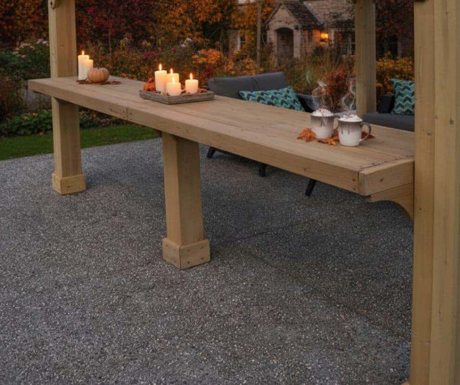 10ft Meridian Gazebo Wooden Shelf 100% Cedar Wooden Shelf Premium Cedar Gazebos with Aluminium Roofs Outdoor wooden shelf under a pergola, styled with lit candles, a small pumpkin, teacups, and scattered autumn leaves, with cushioned chairs and a stone house in the background.