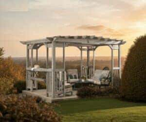 Wooden pergola with a slatted roof, featuring cushioned armchairs and a loveseat arranged around a coffee table, with a side table holding a cake and teapot, set in a landscaped garden with trimmed bushes and a scenic sunset view over distant fields and trees.