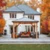 Wooden pergola with string lights and autumn-themed seating area, featuring a sofa and chairs with cushions and throws, set on a stone patio in the landscaped garden of a white house with orange and red trees.