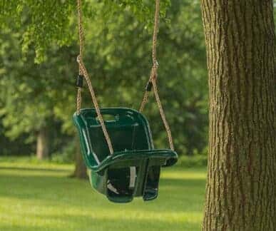 Green plastic baby and toddler swing with a high back and front support, suspended by ropes from a tree branch over a grassy lawn with trees in the background.