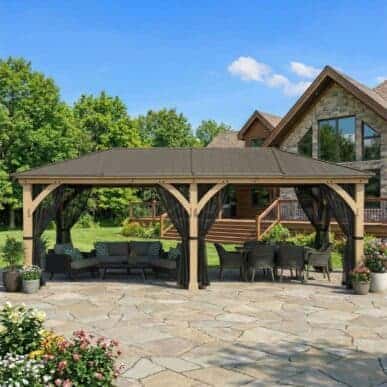 A large wooden gazebo with a dark metal roof and black mesh curtains, featuring an outdoor lounge area with a sectional sofa and a dining table set on a stone patio, surrounded by colourful flowers, landscaping, and a large house in the background.