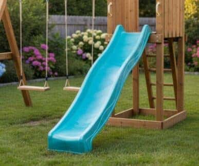 Children’s outdoor playset climbing frame with a green slide, wooden ladder, and two rope swings, set on a grassy lawn with a flower-filled garden and wooden fence in the background.
