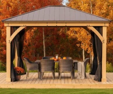 Wooden gazebo with a metal roof and sheer black curtains, set on a deck with a dining table decorated with pumpkins and a candle, surrounded by wicker chairs and autumn foliage.