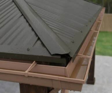 Close-up of a dark corrugated metal roof corner with water droplets and a light brown rain gutter system mounted on wooden beams.