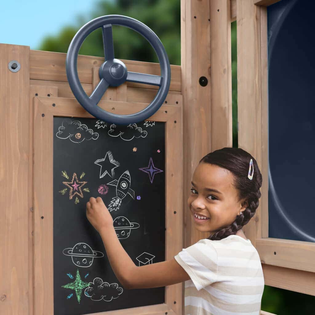 Child drawing space-themed chalk art on a blackboard attached to a wooden play structure with a steering wheel—set in a garden.