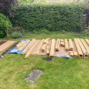Neatly arranged wooden components of various shapes and sizes on a grassy lawn, with curved beams, planks, blocks, and hardware bags, set in a residential garden with hedge and wooden gate.