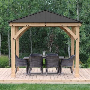 Wooden gazebo with dark metal roof and woven dining set, placed on a wooden deck in a green garden with trees and grass.