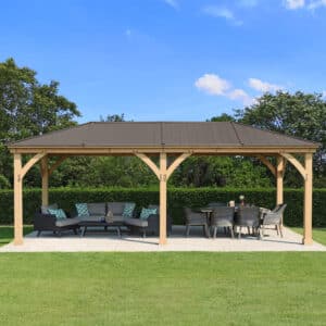 Large wooden gazebo with dark metal roof, featuring a sectional sofa and dining table on a concrete patio in a landscaped garden.