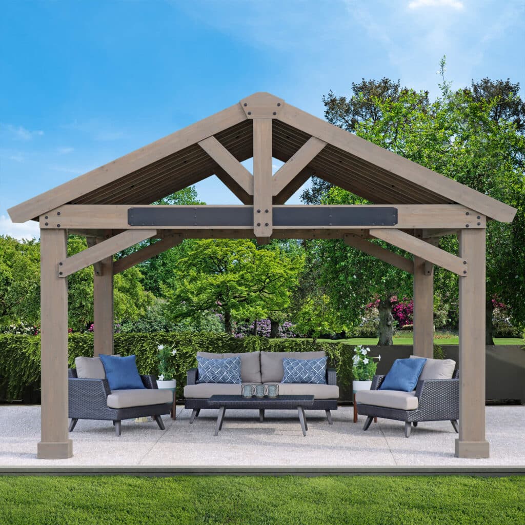 Wooden gazebo with peaked roof and cushioned seating, set on a concrete patio surrounded by greenery and trees.