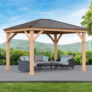 Wooden gazebo with dark metal roof and cushioned seating, set on a paved surface with a scenic backdrop of trees and rolling hills.