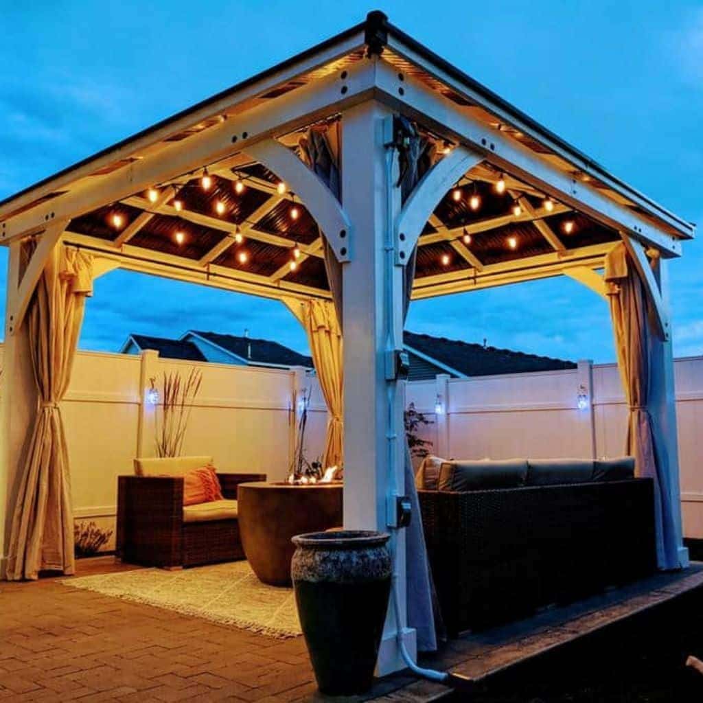 Evening patio setup under a white gazebo with string lights, cushioned wicker furniture, fire pit table, and beige curtains.