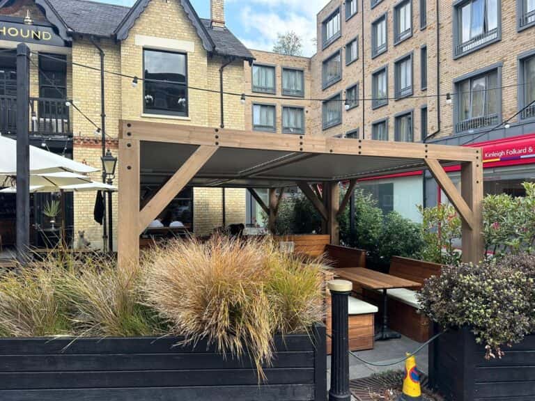 Wooden pergola with string lights over outdoor dining benches in a landscaped urban restaurant setting