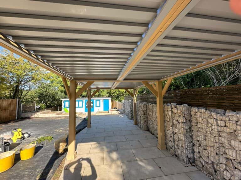 Covered patio with corrugated metal roof, gabion walls, stone paving, yellow planters, and surrounding trees.