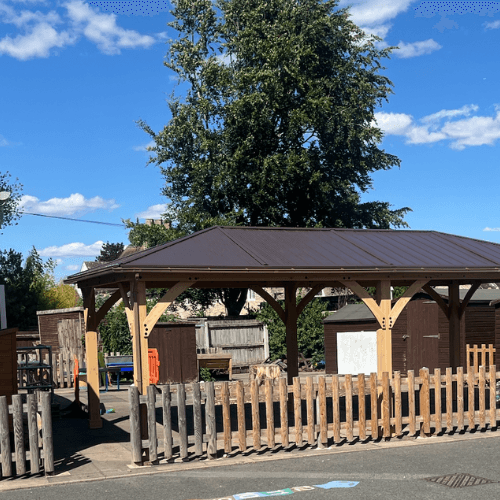 Wooden gazebo with metal roof in fenced school yard with sheds, tree, and playground markings