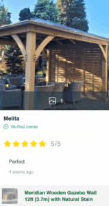 Wooden gazebo with slatted wall, wicker furniture, and pitched roof in garden setting