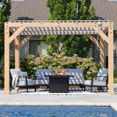 Wooden pergola with slatted roof over cushioned armchairs and fire pit table, set beside a swimming pool with lush garden and hydrangeas in the background.