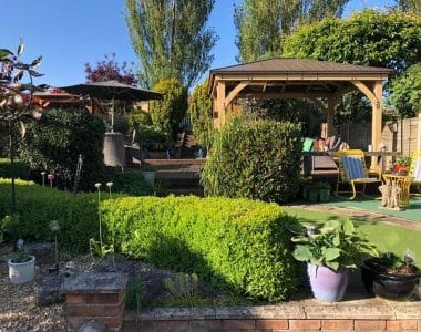 Backyard garden with wooden gazebo, manicured lawn, potted plants, brick planter, and shaded seating areas.