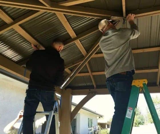 Two people assembling a timber-framed outdoor structure with a corrugated metal roof beside a house