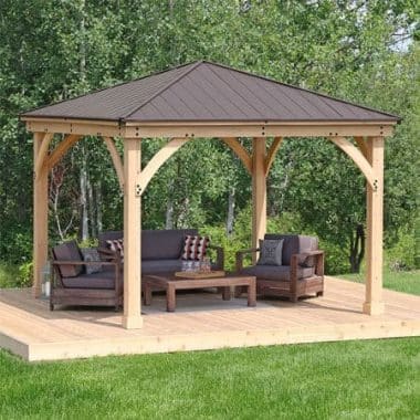 Wooden gazebo with pitched roof and cushioned seating on a raised platform, set in a lush green garden with forest backdrop.