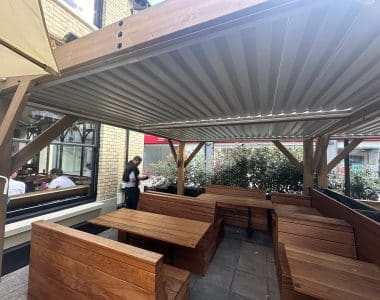 Outdoor dining area with wooden benches and tables beneath a metal-roofed pergola, adjacent to a brick building.