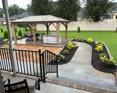 Backyard patio with wooden gazebo and metal roof, dining furniture, outdoor kitchen with stone facade, brick paving, flower beds, and white privacy fence.